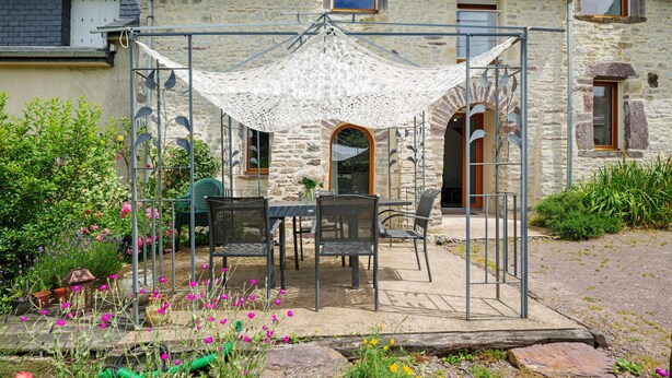 A stone building with a covered patio area featuring a table and chairs, surrounded by greenery and flowers.