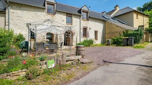 Une maison en pierre dotée d’un espace extérieur couvert avec des sièges, d’un jardin fleuri et d’une allée en gravier.