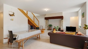 Een woonkamer met een bank, een televisie, een trap, een eettafel met stoelen en een open haard.