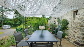 Een overdekt terras met een tafel en stoelen, omringd door groen en een stenen muur.