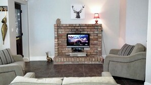 Een woonkamer met een metselwerk open haard, een televisie, twee banken en een decoratief wandkunstwerk.