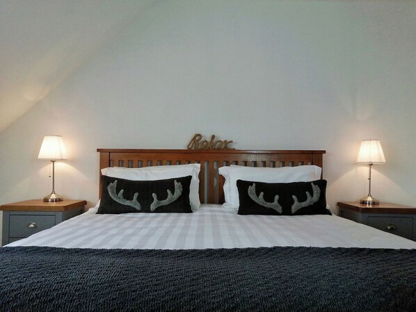 Un lit recouvert d’une couvre-lit à rayures, deux oreillers noirs ornés de motifs de bois de cerf, une tête de lit en bois sur laquelle est inscrit le mot « Relax », et deux lampes de chevet posées sur des tables de nuit en bois.