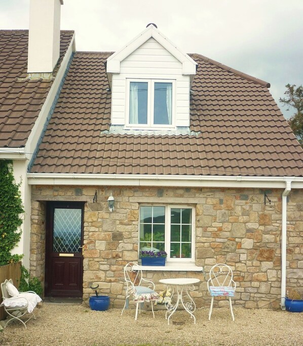 A stone house with a tiled roof, a front door, and a small patio with two chairs and a table.