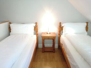 Un petit logement composé d’une seule pièce, comprenant deux lits simples, une table de chevet en bois et une petite table de nuit avec une lampe.