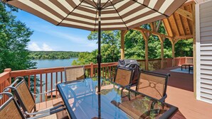 A deck with a glass table and chairs, a striped umbrella, and a view of a lake. - The Lake Escape, Sleeps 8, Big Deep Water, Kayaks, Lily Pad + MORE! (Sparta)