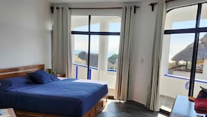 A bedroom with a bed, a ceiling fan, a wooden dresser, and a view of the ocean through large windows.