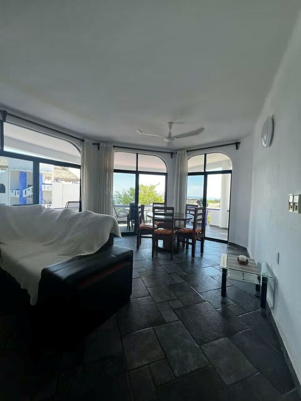 A hotel room with a bed, a dining table, chairs, a ceiling fan, and large windows offering a view.