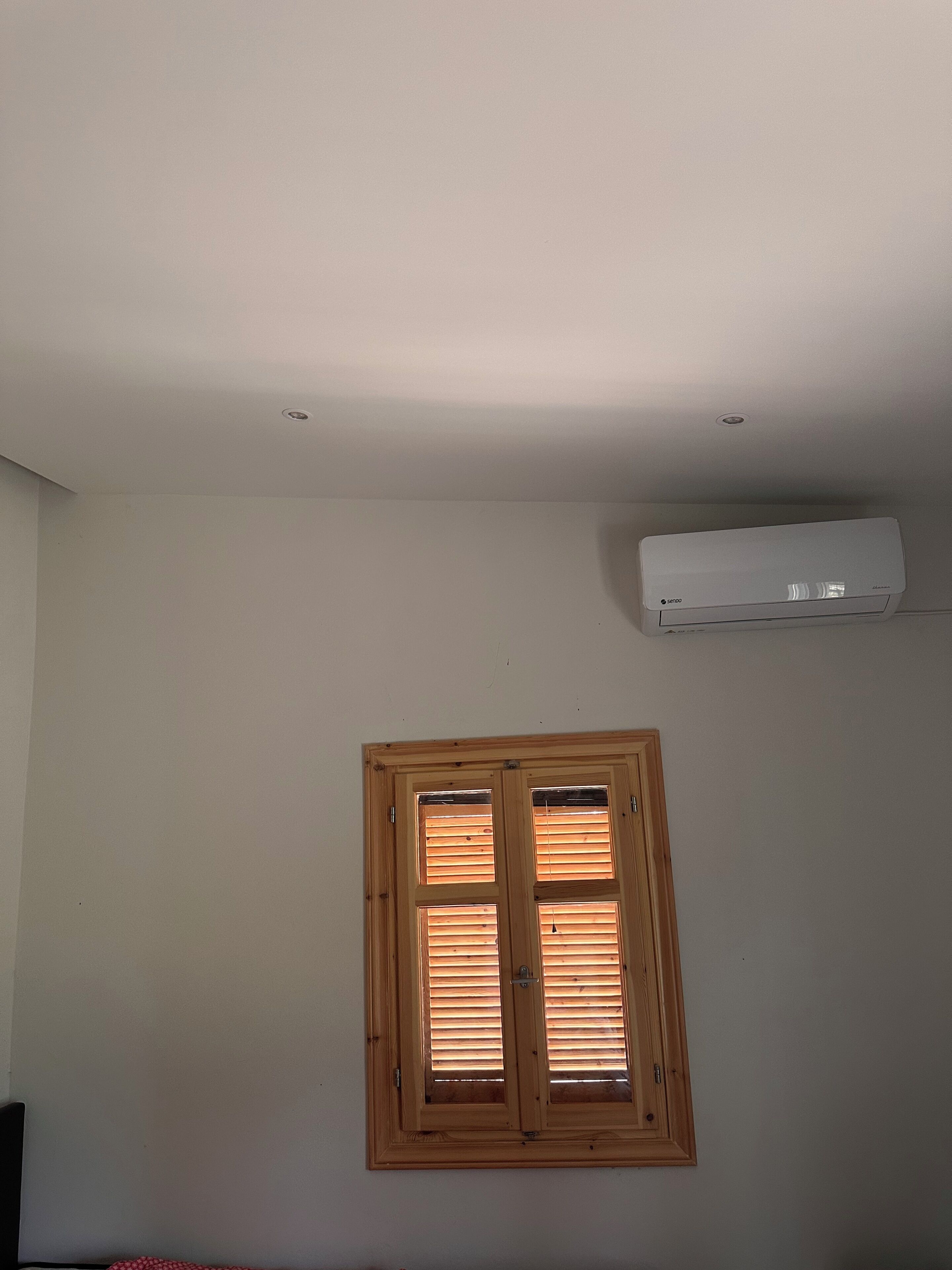 Quarto com janela emoldurada em madeira, ar condicionado fixado na parede e teto com iluminação embutida.