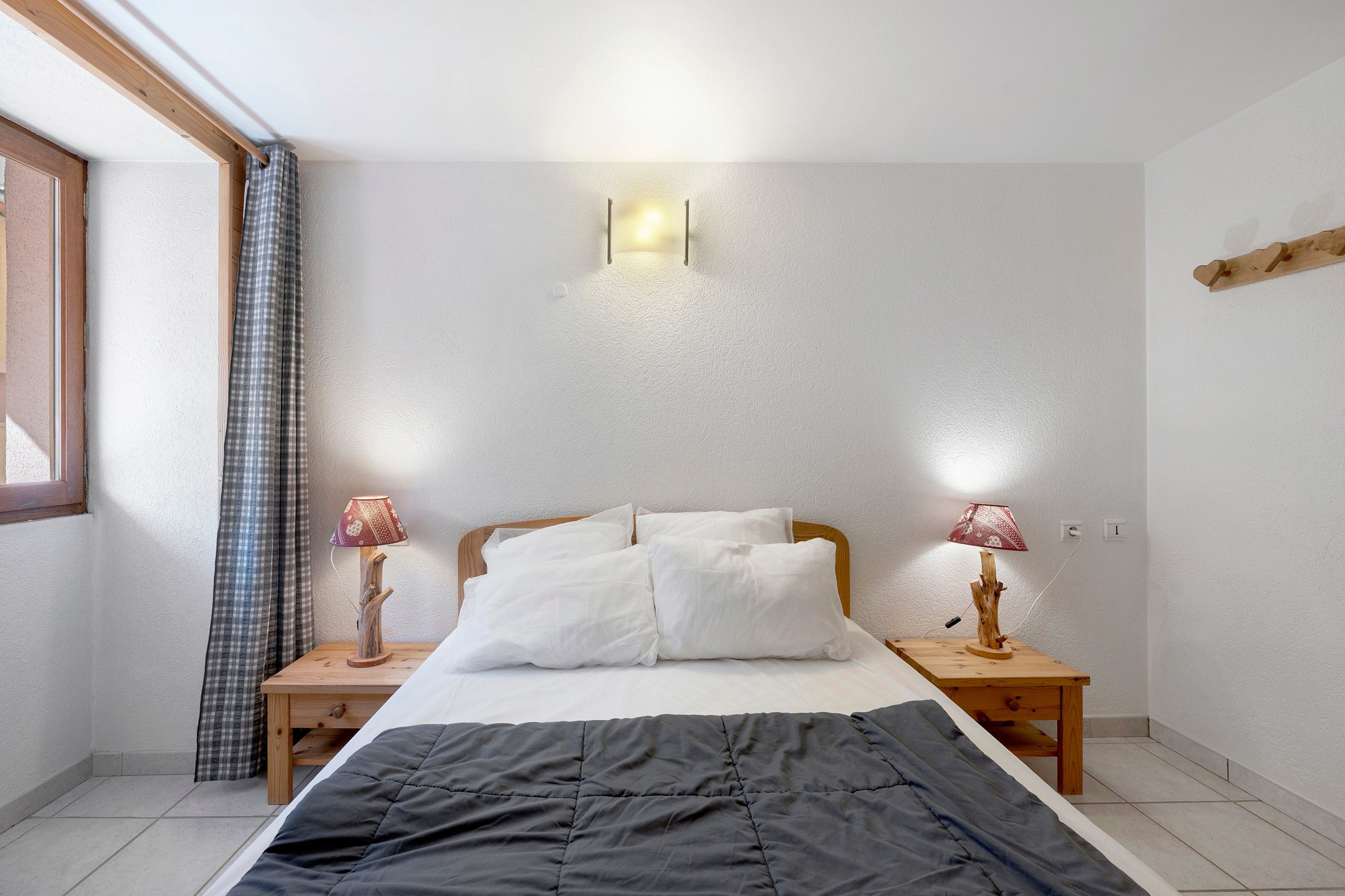 Un dormitorio con una cama, dos mesitas de noche con lámparas, una ventana con cortinas y una lámpara de pared.