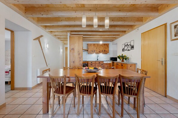 Un comedor con mesa y sillas de madera, una cocina al fondo y techo de madera.