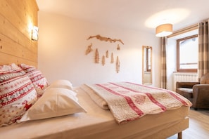 Ein Schlafzimmer mit Bett, Kissen, einer Steudecke, Holzvertäfelung, Wandleuchten, einem Spiegel, einem Fenster mit Vorhängen und einem Sessel.