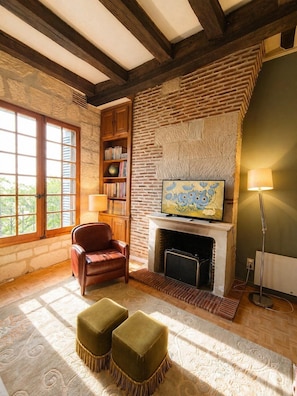 A cozy living room with a fireplace, a leather armchair, ottomans, a bookshelf, and a painting on the wall.
