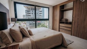 Ein Schlafzimmer mit großem Fenster, einem Bett mit beige farben gewählter Bettwäsche, einem hölzernen Kleiderschrank und Blick auf die äußere Bebauung.