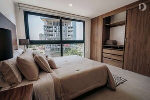 Ein Schlafzimmer mit großem Fenster, einem Bett mit beige farben gewählter Bettwäsche, einem hölzernen Kleiderschrank und Blick auf die äußere Bebauung.