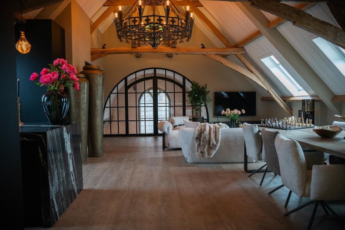 Um loft espaçoso com teto alto, um lustre, área de jantar com um tabuleiro de xadrez e uma grande janela em arco.