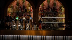 A bar with two arched windows, shelves of liquor bottles, and a bar counter with taps and glasses. - Room4 (Maurik)