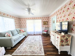 A room with a ceiling fan, floral wallpaper, a sofa, a rocking chair, a television, and a patterned rug.