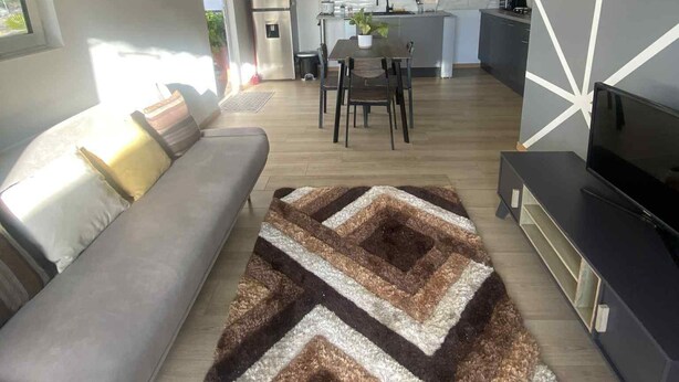 A modern living room with a sofa, a rug, a TV, and a kitchen in the background. - Bungalow Le Sapotille (Saint-Paul)