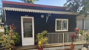 A small house with a dark exterior, a white door, and a small window. There are potted plants and a white railing along the front porch. - Bungalow Le Sapotille (Saint-Paul)