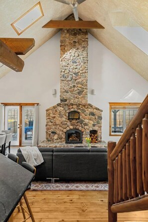 A cozy living room with a stone fireplace, wooden ceiling beams, and a view through a glass door.