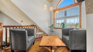 Una habitación estilo loft con una escalera de madera, dos sofás grises, una mesa de madera y un ventanal con persianas.