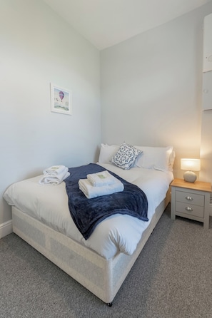 A neatly made bed with white linens, a blue throw blanket, and a decorative pillow. There is a small bedside table with a lamp and a framed picture on the wall.