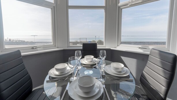 A dining table set for six with white plates, glasses, and cutlery, positioned in a room with large windows overlooking a body of water. - Cosy 2-Bed apartment | sleeps 6 | Sea views - Pass the Keys (Southend-on-Sea)