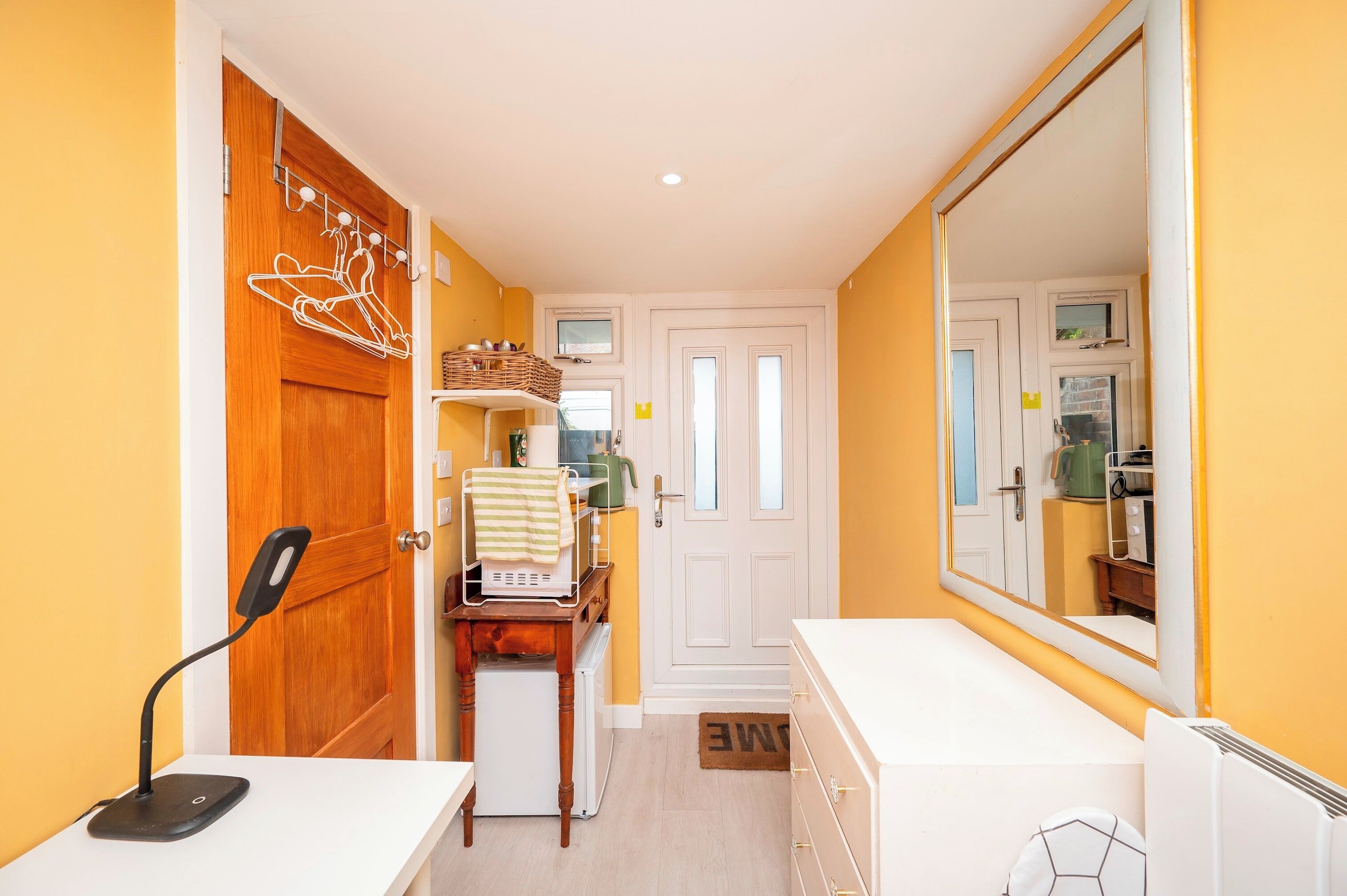 A narrow hallway with a wooden door, a white door, and a mirror reflecting a kitchen area.
