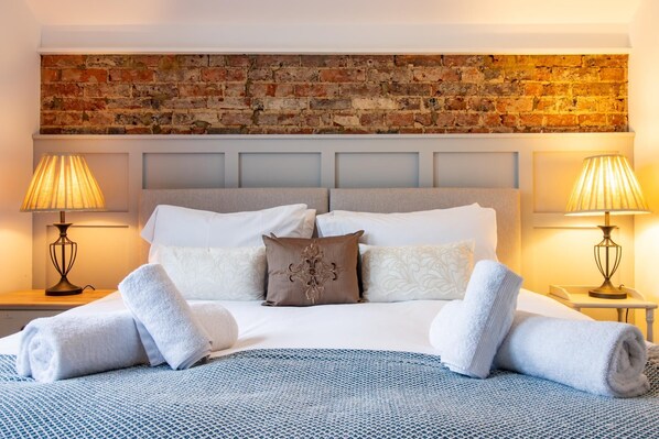 A neatly made bed with white linens and rolled towels, a brick wall behind, and two bedside lamps on either side.