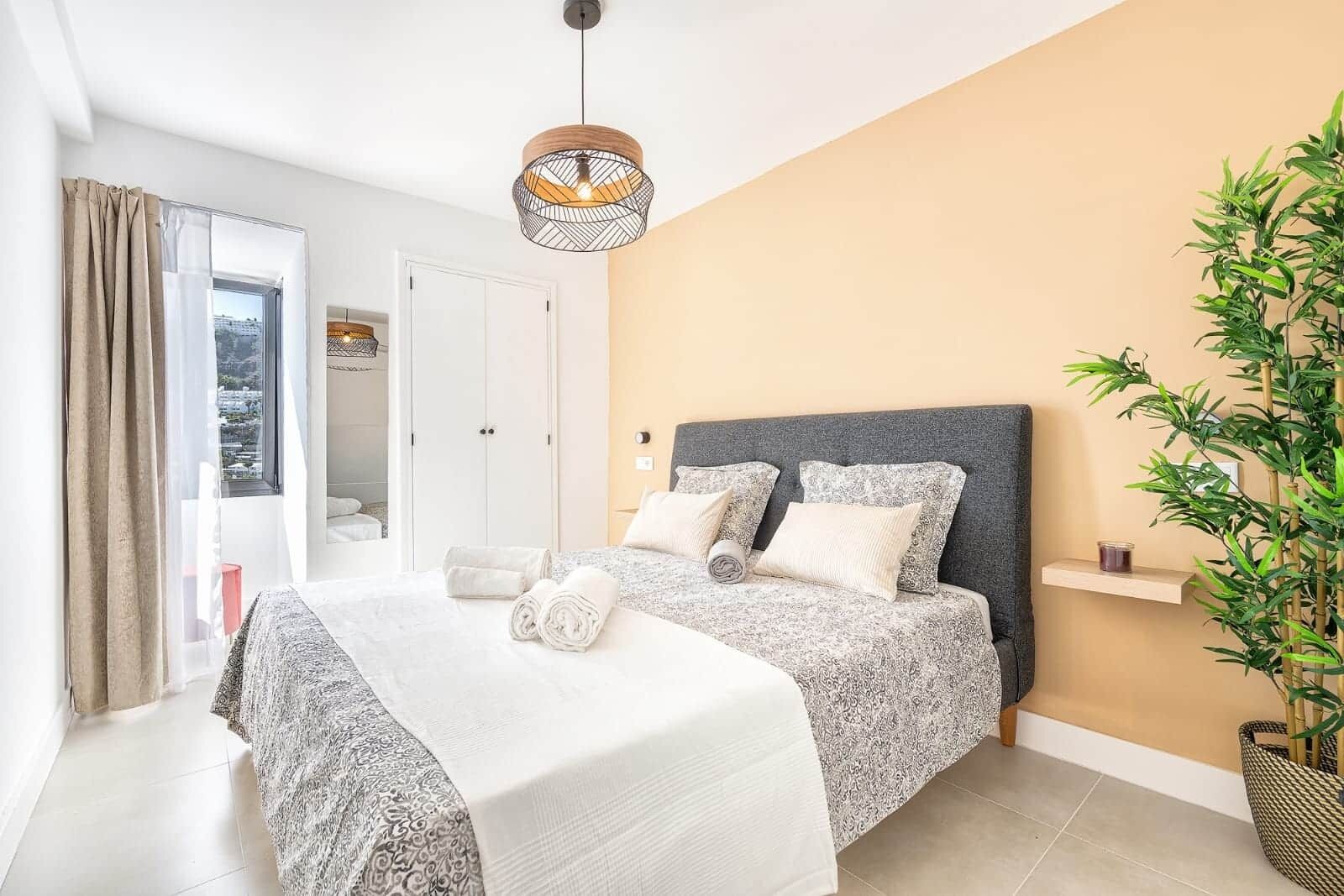 Ein Schlafzimmer mit einem Bett, Kissen, einer Hängeleuchte, einer Tür und einer Pflanze.
