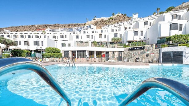A swimming pool with clear water, surrounded by white buildings with balconies, and a rocky hillside in the background. - Casa Arizona – Central Duplex Puerto Rico GC (Puerto Rico de Gran Canaria)