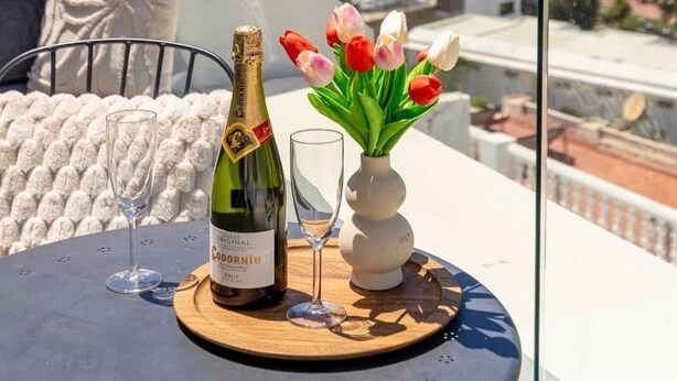 Un balcón con una mesa dispuesta para una bebida, un jarrón de tulipanes y vistas al mar y edificios.