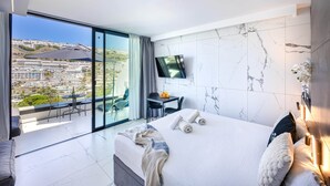 Una habitación de hotel moderna con una cama grande, un televisor montado en la pared, un balcón con mesa y sillas, y vistas a la ciudad.