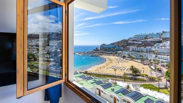 Vista costeira por uma janela, com uma praia, um porto de iates e uma colina de edifícios.