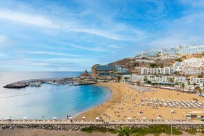 A coastal town with a beach, a rocky pier, and buildings with balconies overlooking the sea. - Modern Sea View Apartment | Pool | Walk to Beach | Puerto Rico (Puerto Rico de Gran Canaria)