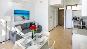 Uma sala de estar moderna com uma mesa de jantar posta para quatro pessoas, um sofá cinza com um almofada, um quadro de um barco na água e uma área de cozinha com geladeira e fogão.