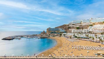Un pueblo costero con una playa, un muelle rocoso y edificios con balcones que dan al mar.