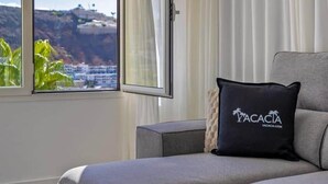 Ein modernes Wohnzimmer mit einer grauen Eckcouch, einem kleinen Beistelltisch und einem schwarzen Kissen mit dem Schriftzug 'VACACIA'. Ein großes Fenster bietet Blick auf eine Küstenlandschaft.