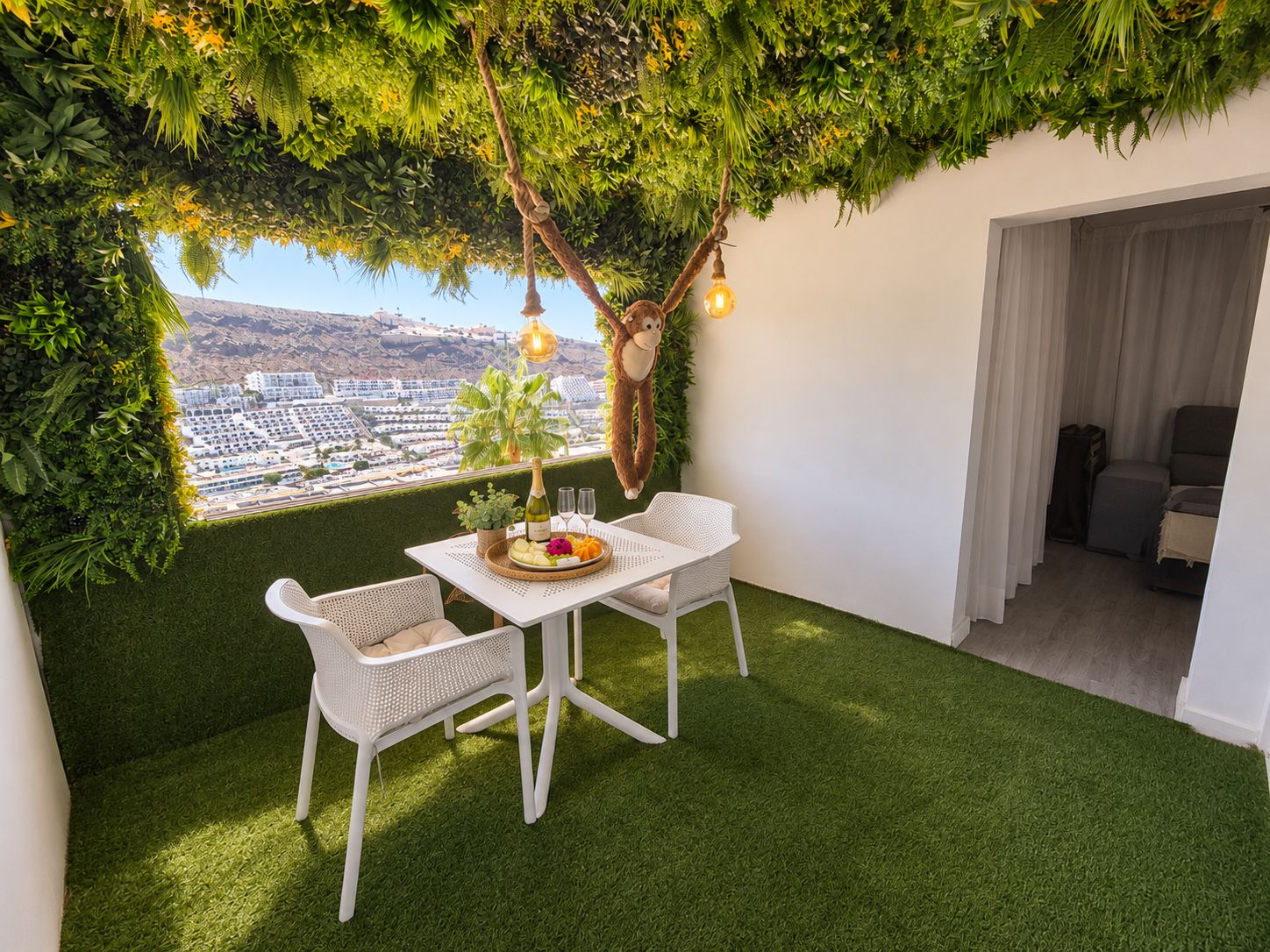 A balcony with artificial grass, white wicker furniture, a dining table with a bottle and glasses, and a view of a cityscape.
