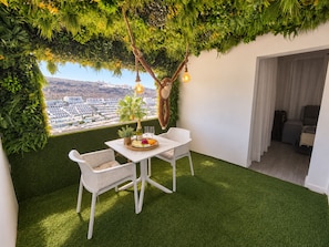 A balcony with artificial grass, white wicker furniture, a dining table with a bottle and glasses, and a view of a cityscape. - Sea View Apartment with Pool – Canaima, Puerto Rico (Puerto Rico de Gran Canaria)