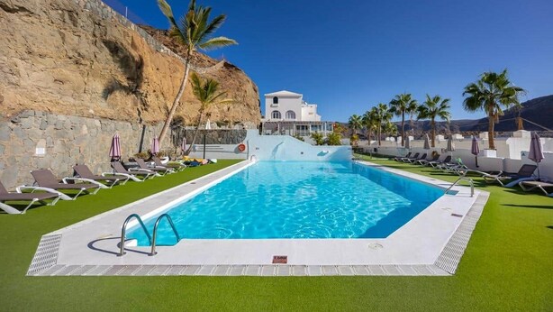 Ein Pool mit Liegestühlen und Palmen, umgeben von einer Steinmauer und einem Gebäude im Hintergrund.