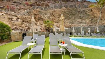 Zona de piscina con sillones de descanso y sombrillas, piscina y colina rocosa al fondo.