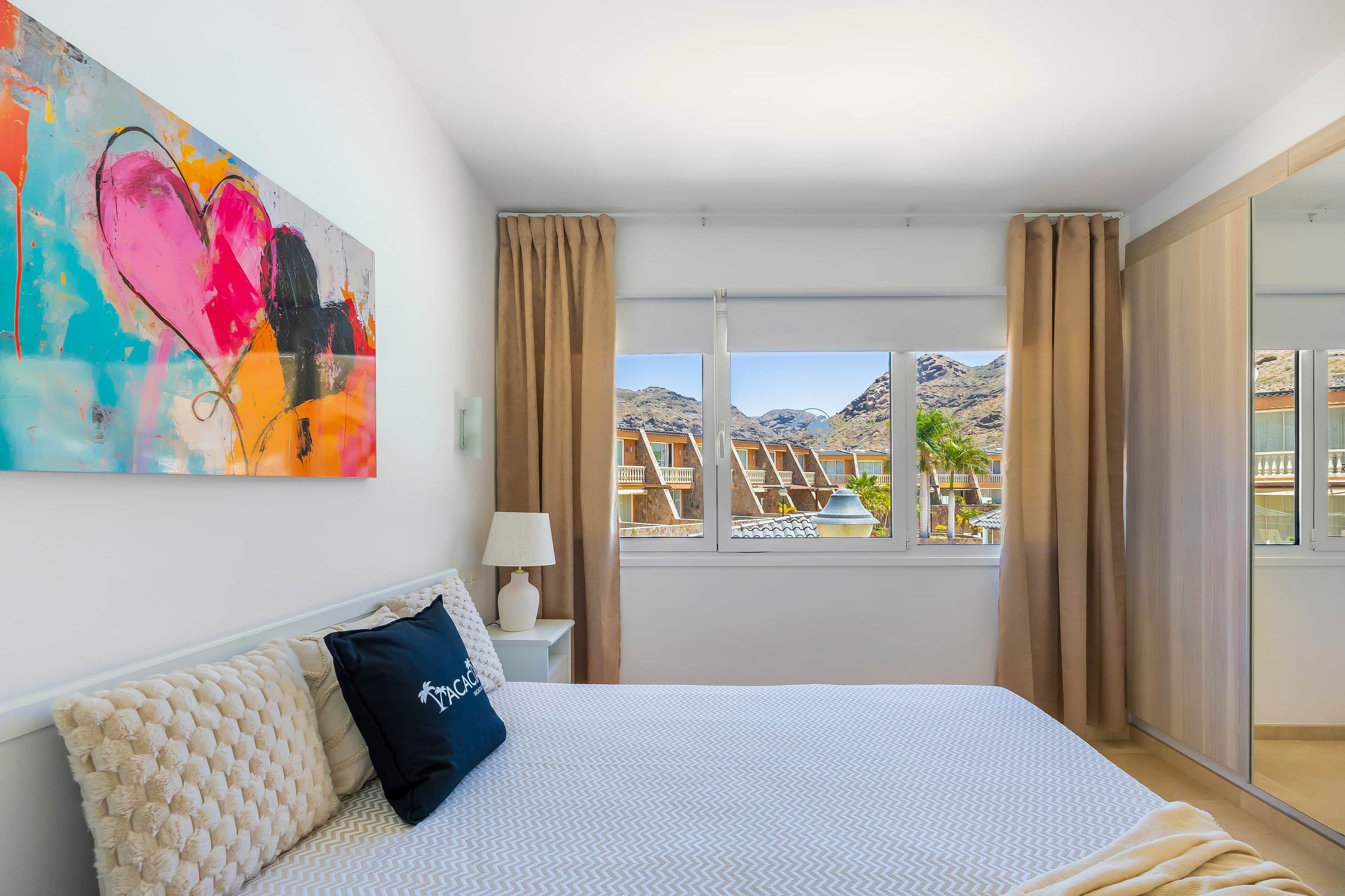 Habitación de hotel con cama, un cuadro, una ventana con cortinas y vistas a edificios y montañas.