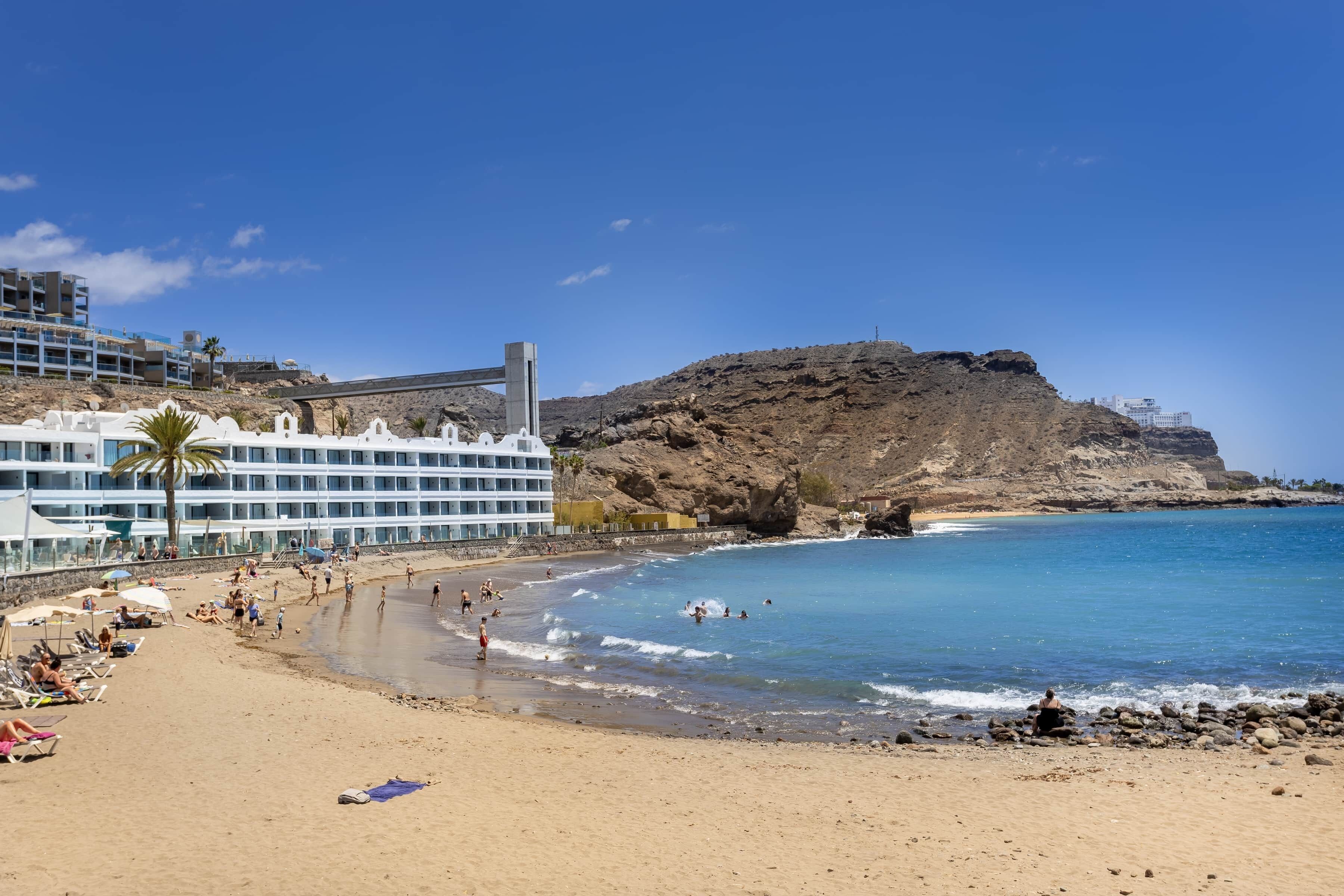 A beach with a hotel, rocky cliffs, and clear blue water.
