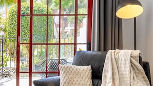 Un salon moderne avec un canapé en cuir foncé, un plaid blanc à texture, une lampe de sol noire et une vue, à travers une porte vitrée à cadre rouge, sur un espace extérieur verdoyant.