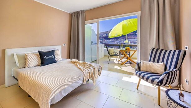 Una habitació d'hotel amb un llit, un butac armchair rayat, una taula petita amb una ampolla i un balcó amb un para-sol groc.