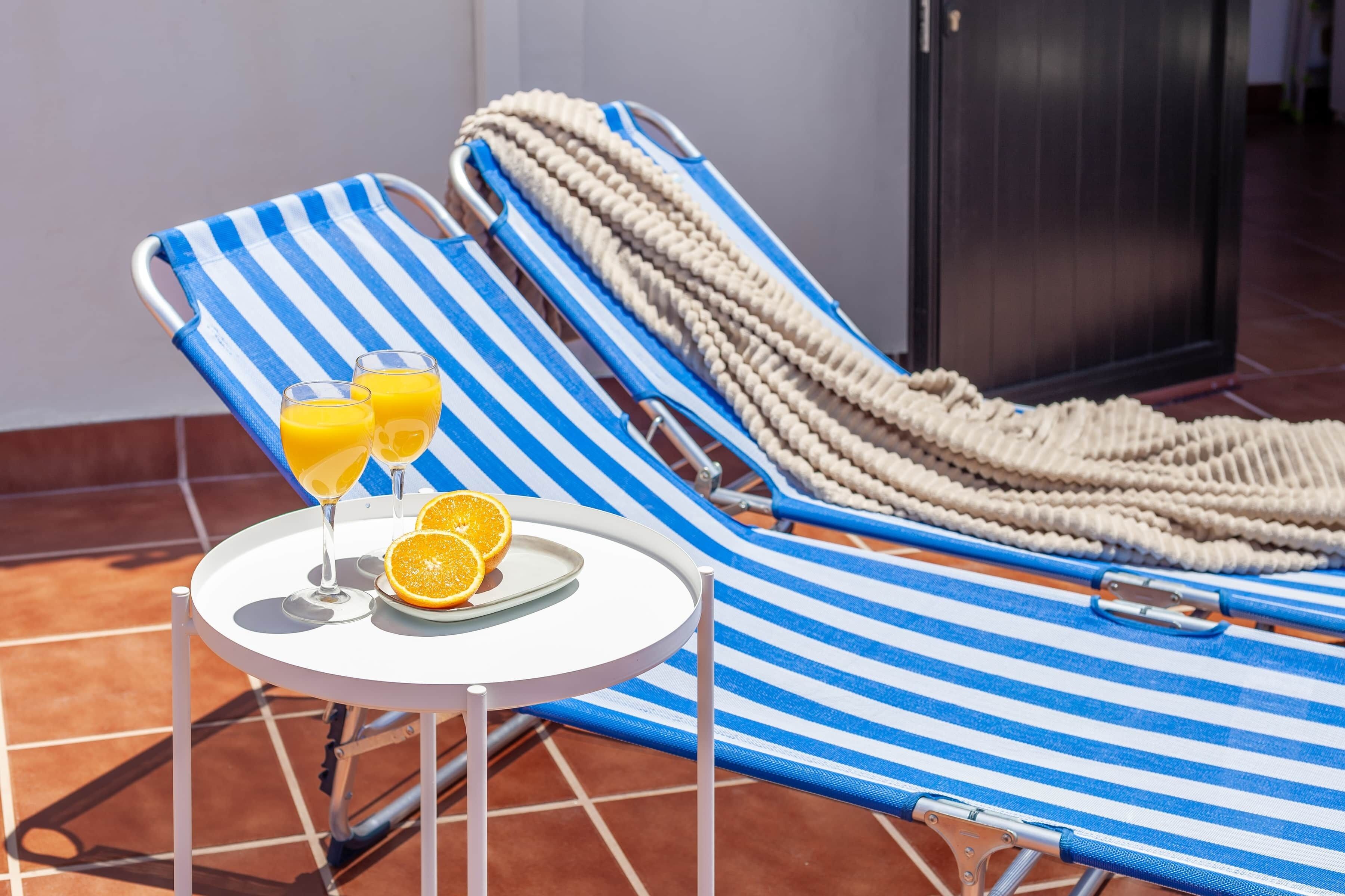 Una cadira de sol amb teixit blau i blanc a ratlles, una taula rodona petita amb un plat de menjar i dos vasos de suc d'aranja, i una corda beix per donar suport.