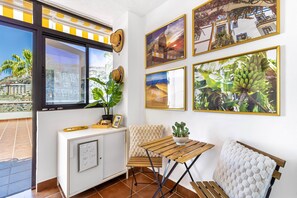Una habitación con ventana, un armario blanco, un banco acolchado, una mesa de madera y un cuadro en la pared.