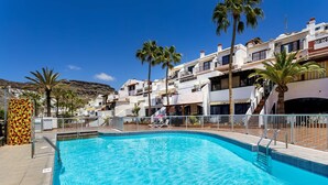 Une piscine à l’eau d’un bleu cristallin, entourée de bâtiments blancs dotés de balcons, de palmiers et surplombée par un paysage montagneux.