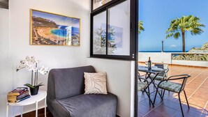 Une pièce avec un canapé gris, une petite table ronde entourée de chaises, une orchidée en pot, un tableau encadré représentant une plage, et une grande fenêtre donnant sur un patio avec vue sur la mer et les palmiers.